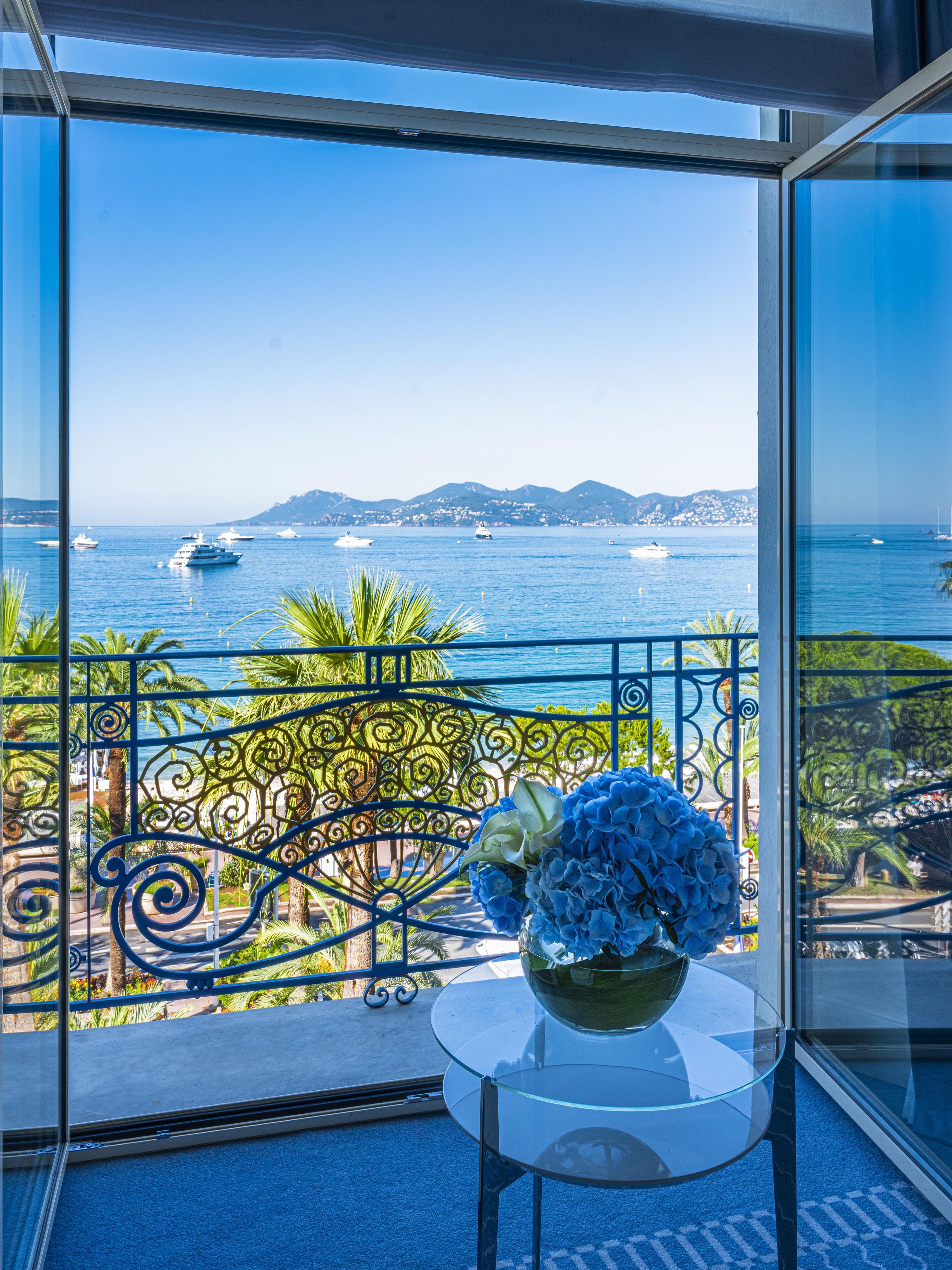 Un balcon avec une table en verre et un vase de fleurs, offrant une vue sur la côte, avec des bateaux et des montagnes en toile de fond.