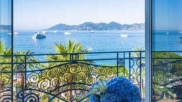 Un balcon avec une table en verre et un vase de fleurs, offrant une vue sur la côte, avec des bateaux et des montagnes en toile de fond.