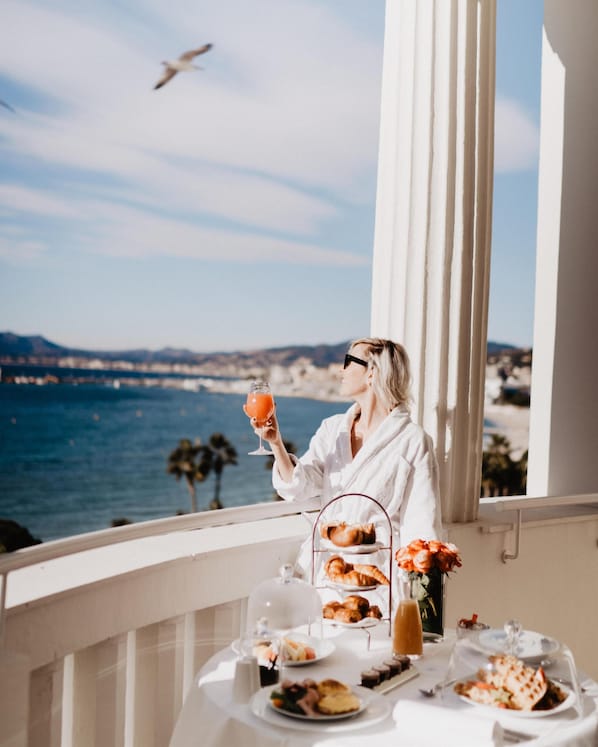 A balcony with a table set for breakfast, overlooking a coastal view with the sea and mountains in the background.