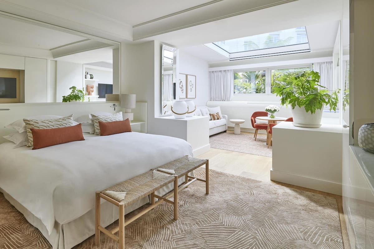 Une chambre d’hôtel moderne avec un grand lit, une banquette, un bureau et une chaise. On y trouve également un grand miroir, une fenêtre donnant sur l’extérieur et une lucarne.