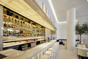 A modern bar area with a long counter, multiple shelves of bottles, and a seating area with white chairs and tables.