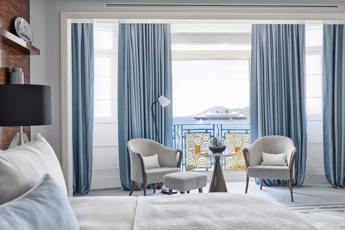 Une chambre d’hôtel avec un lit, deux fauteuils, une petite table et une vue sur la mer.