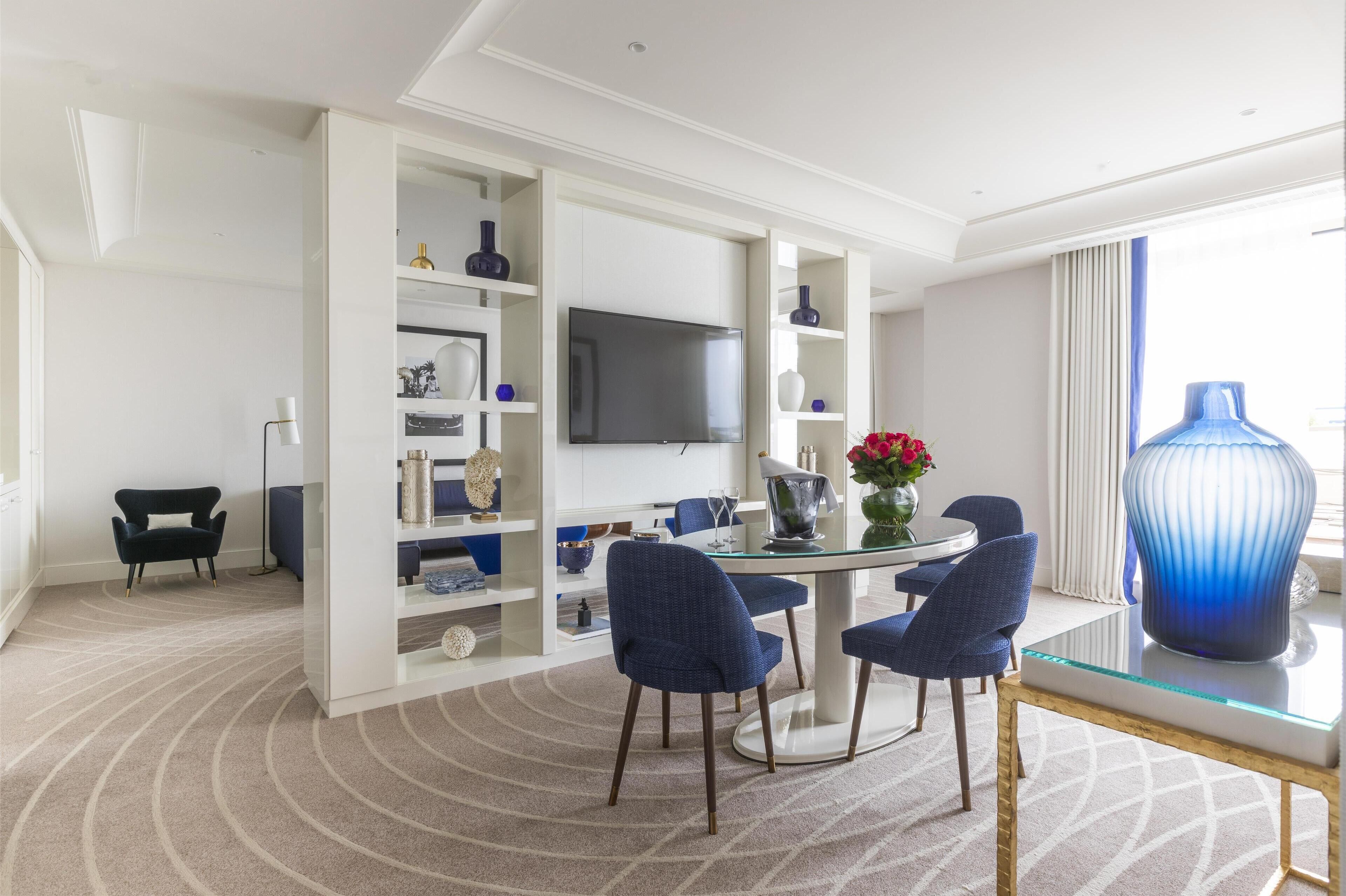 A modern hotel room with a round dining table, blue upholstered chairs, a large blue vase, and a flat-screen TV mounted on the wall.
