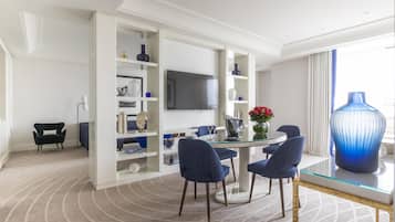 A modern hotel room with a round dining table, blue upholstered chairs, a large blue vase, and a flat-screen TV mounted on the wall.