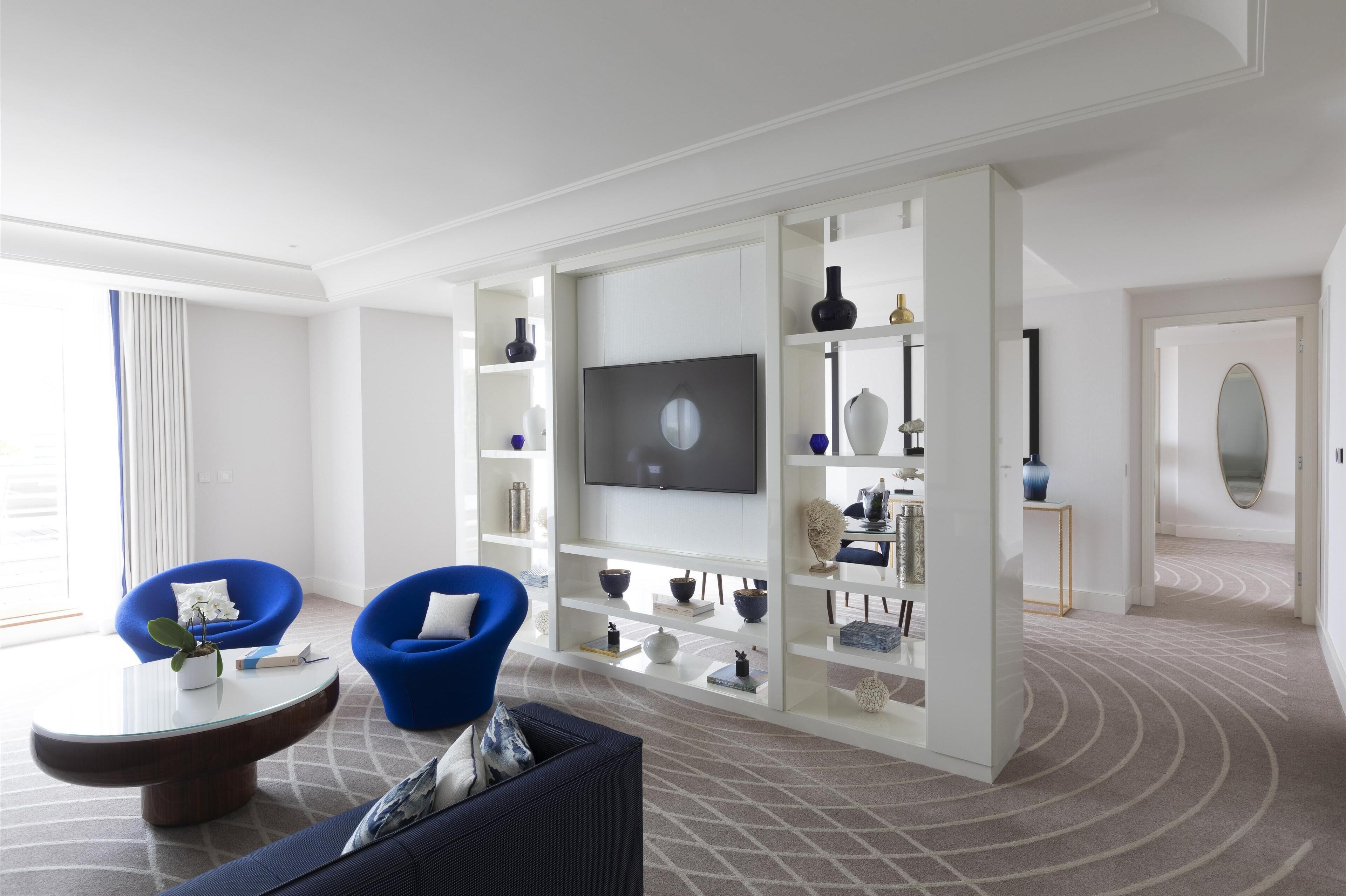 Un salon moderne doté d’un téléviseur à écran plat, de fauteuils bleus, d’une table basse et d’un meuble de rangement blanc avec des étagères.