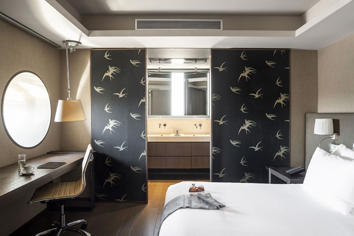 A hotel room with a bed, a desk with a lamp, a round mirror, a bathroom with a sink and mirror, and a window.