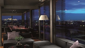 A modern living room with a city view, a dining area with red table settings, and a large window with blinds. - Radisson Blu Park Hotel, Athens (Athens)