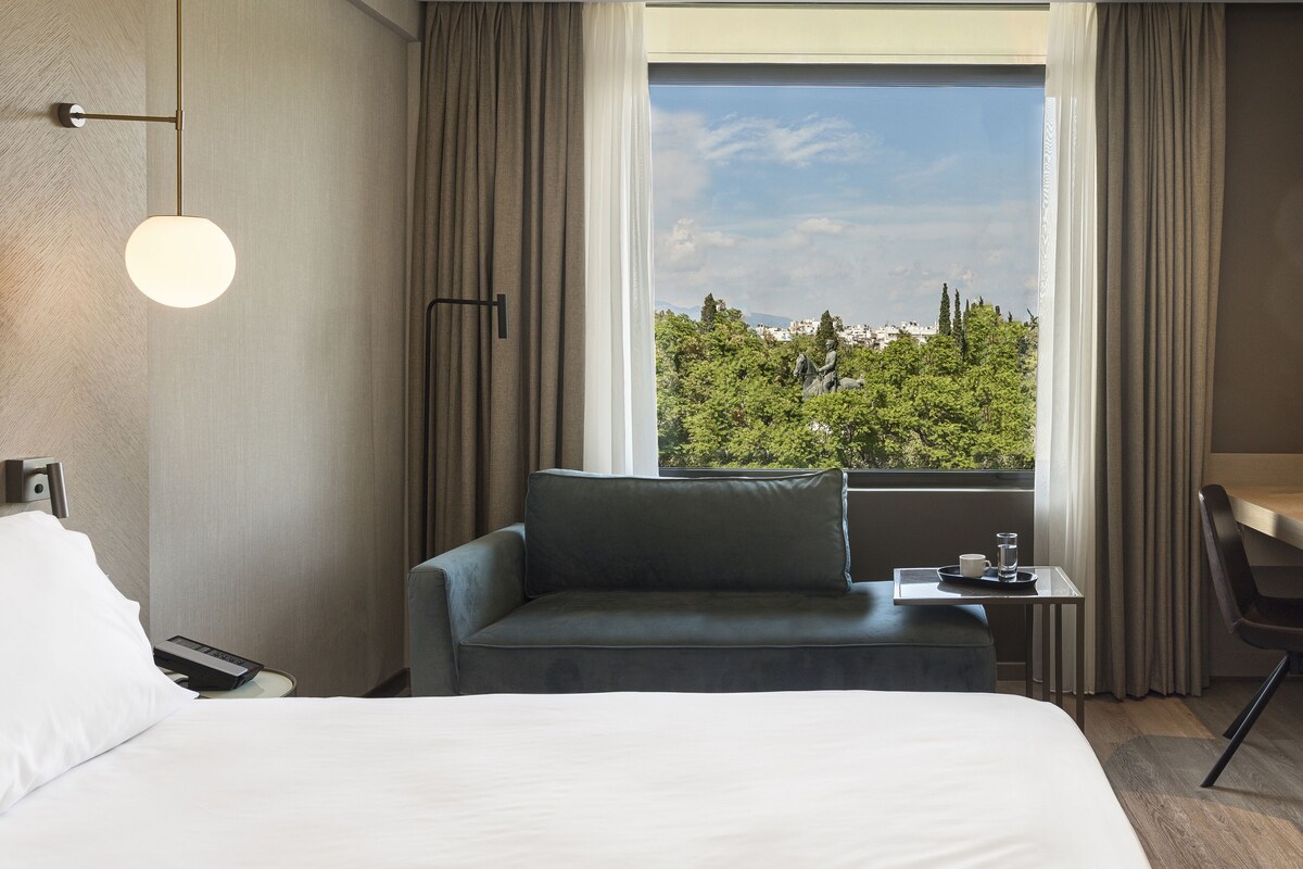 A hotel room with a bed, a sofa, a desk, a lamp, and a view of a cityscape through large windows.