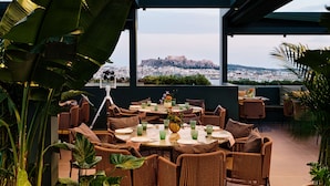 An outdoor dining area with wicker chairs and tables, large potted plants, and a view of a cityscape with a historic building. - Radisson Blu Park Hotel, Athens (Athens)