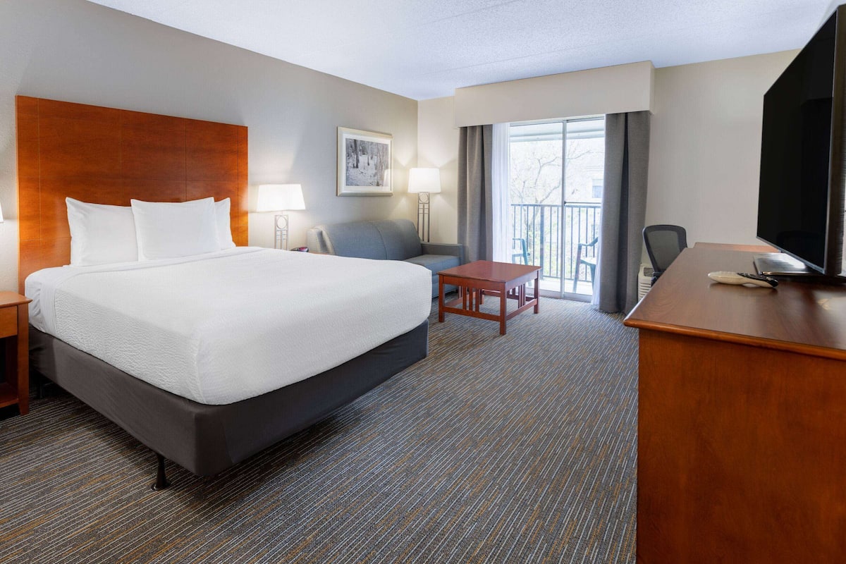 A hotel room with a large bed, a desk, a chair, a TV, and a balcony with a view.