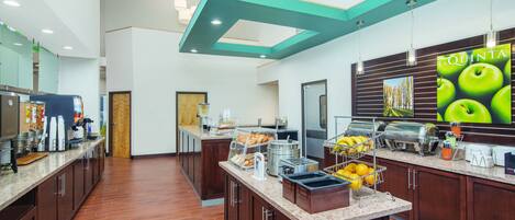 A modern hotel kitchen with a breakfast bar, coffee machines, and a large poster advertising 'La Quinta'.