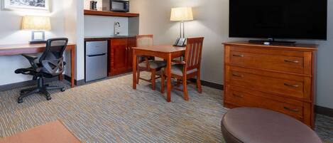 Una habitación de hotel con kitchenette, un comedor con mesa y sillas, un televisor, un escritorio con silla y un pequeño taburete.