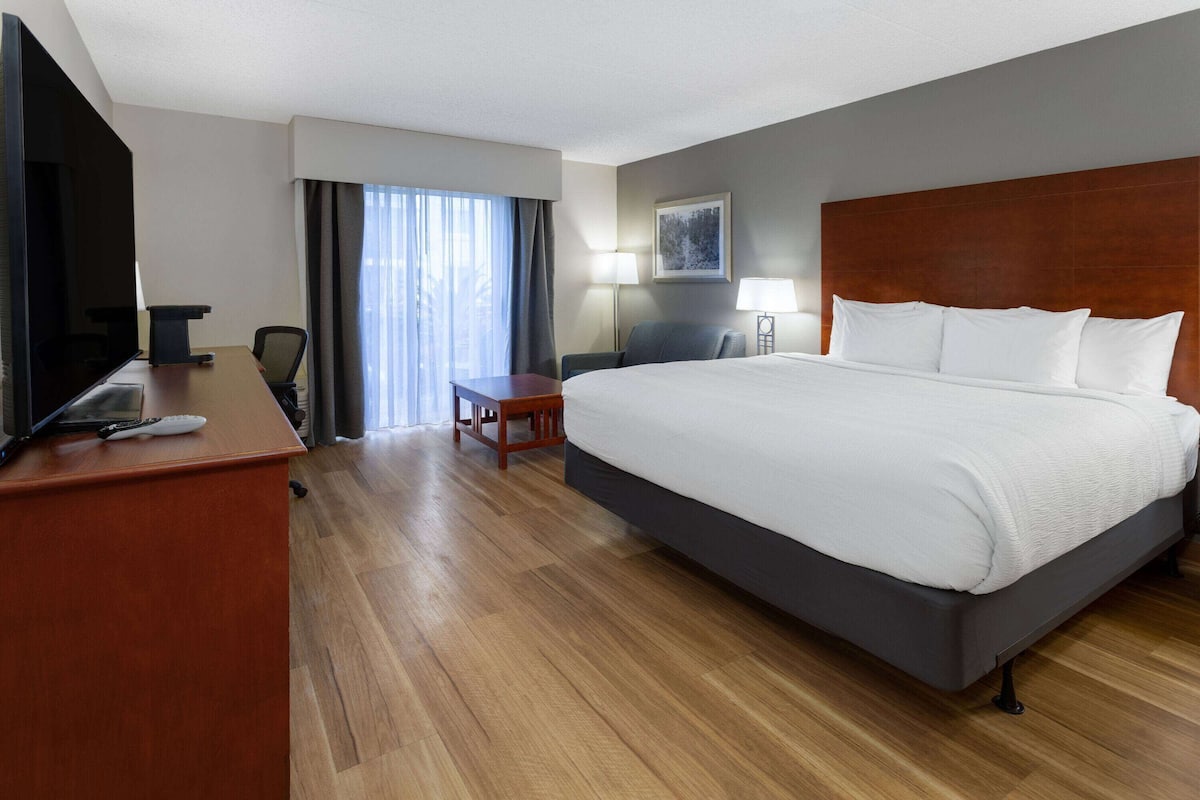 A hotel room with a large bed, a desk with a TV, a chair, a small table, and a window with curtains.
