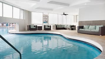 An indoor swimming pool with a sunlit room featuring a sofa and a ceiling fan.