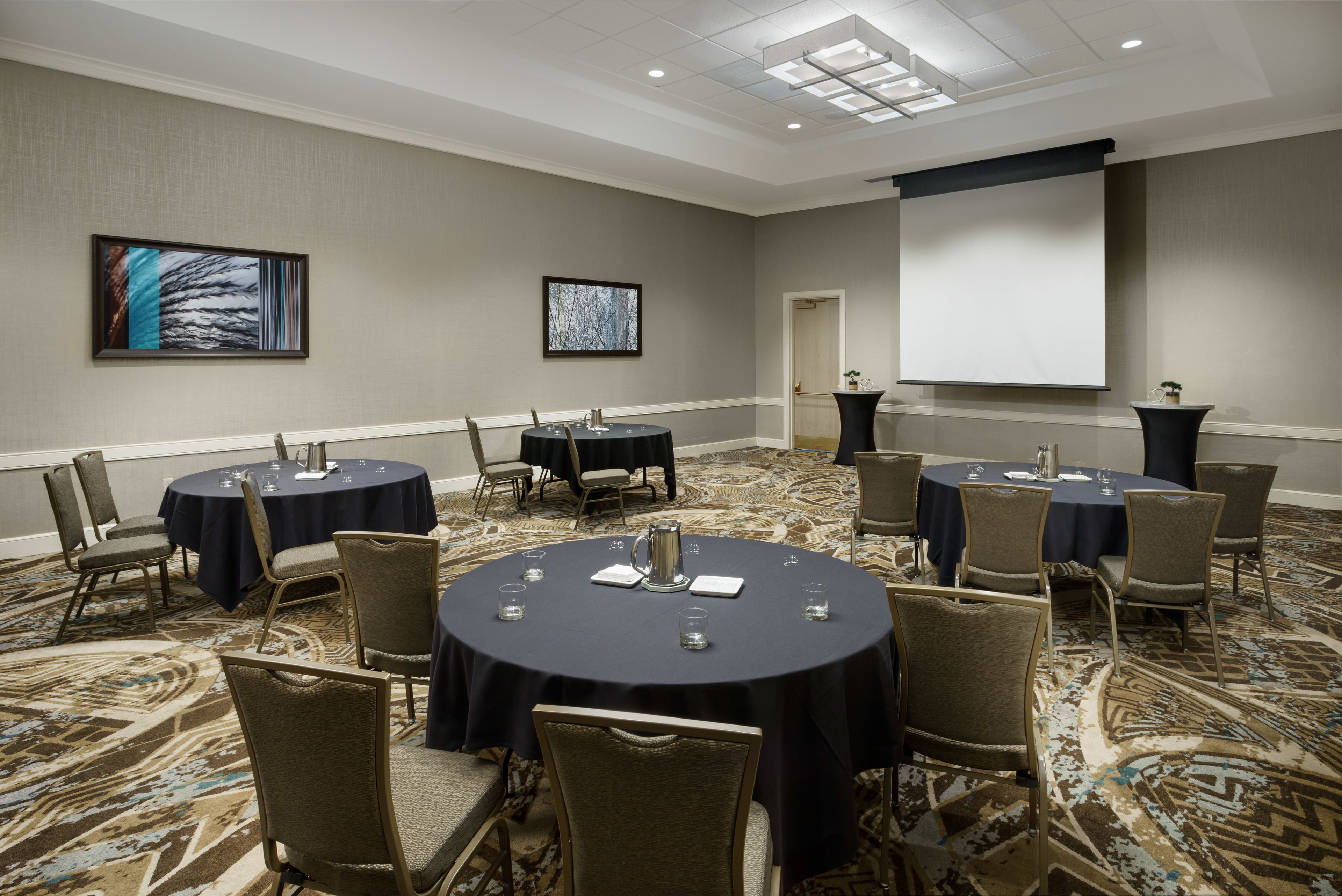 A conference room with round tables and chairs, a projector screen, and two framed artworks on the walls.