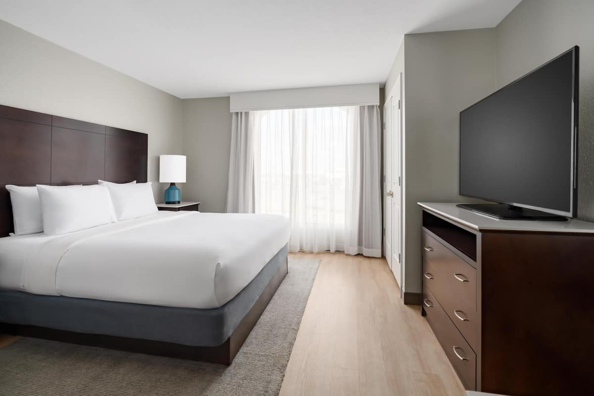 Una habitación de hotel con una cama grande, un televisor en una cómoda, una mesita de noche con lámpara y una ventana con cortinas.