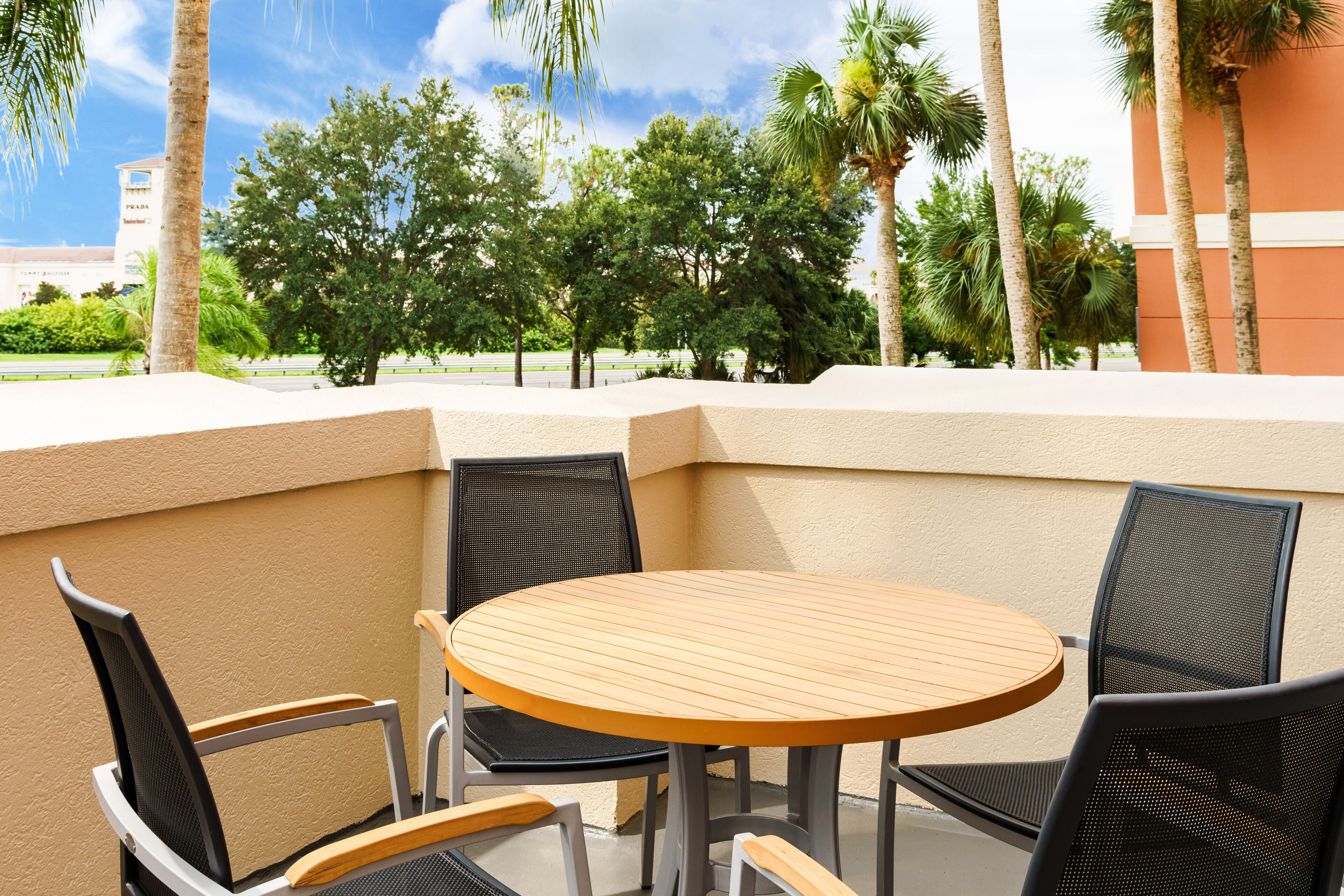 A corner outdoor seating area with a round wooden table and four chairs, surrounded by a beige wall and palm trees.