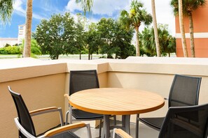Un espacio exterior con mesas y sillas, una mesa redonda de madera y cuatro sillas, junto a un muro color arena y palmeras.