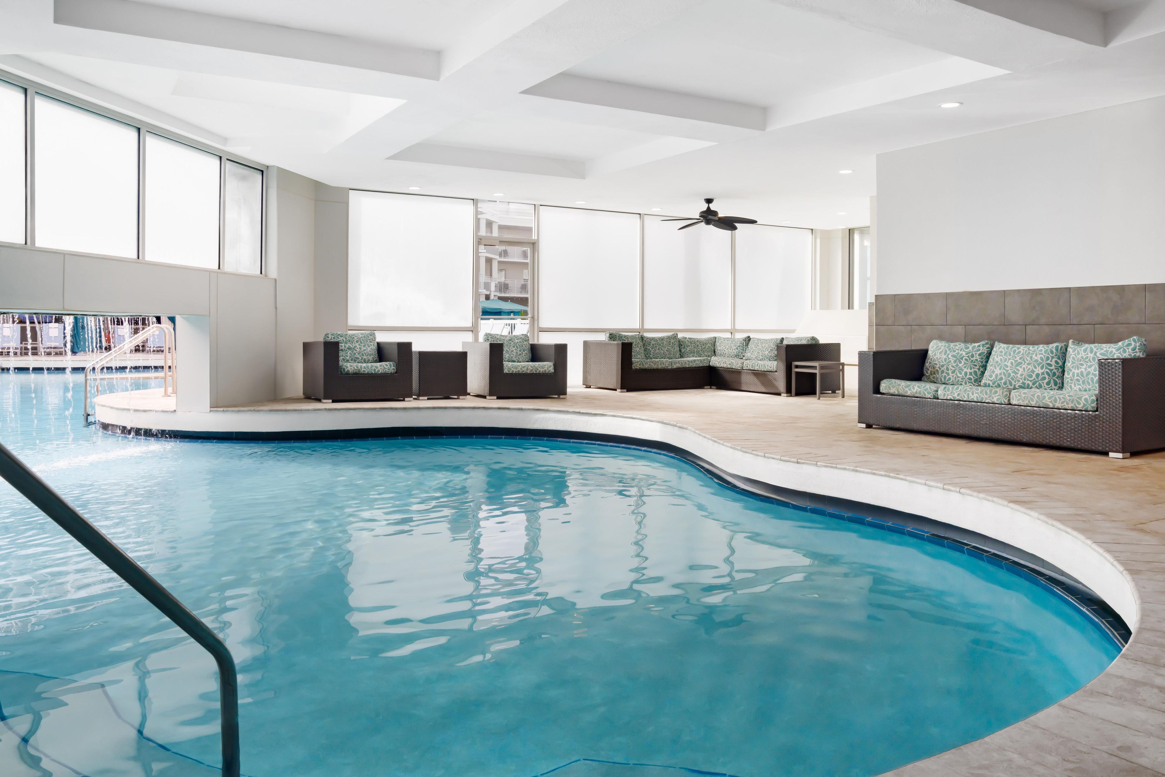 Un espace piscine intérieur moderne, doté d’une piscine aux lignes courbes, de coins salon et d’un ventilateur de plafond.