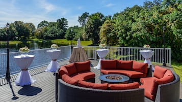A deck with wicker furniture, white tables with flower arrangements, and a view of a lake.