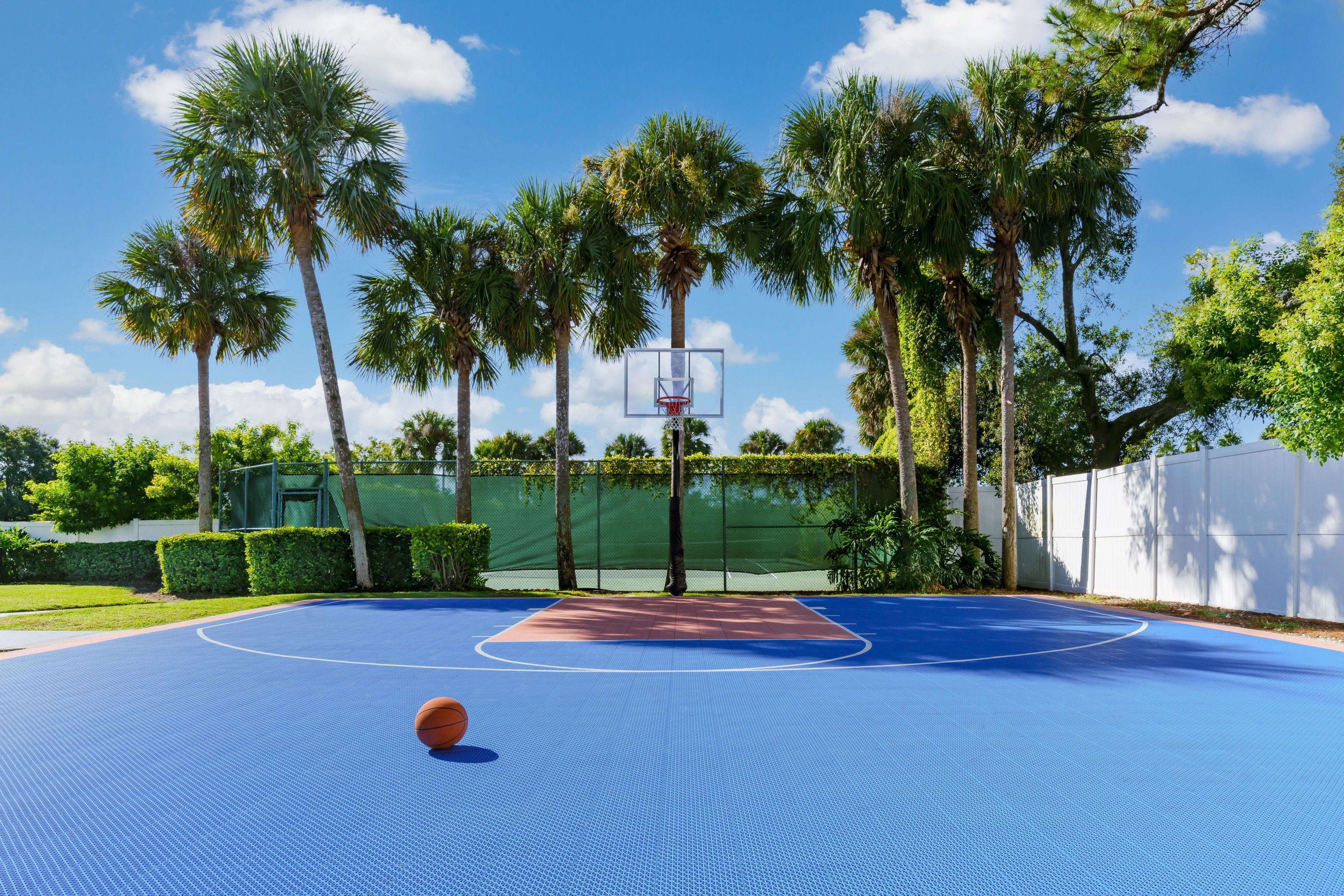 An outdoor basketball court with a blue surface, surrounded by palm trees and a white fence.