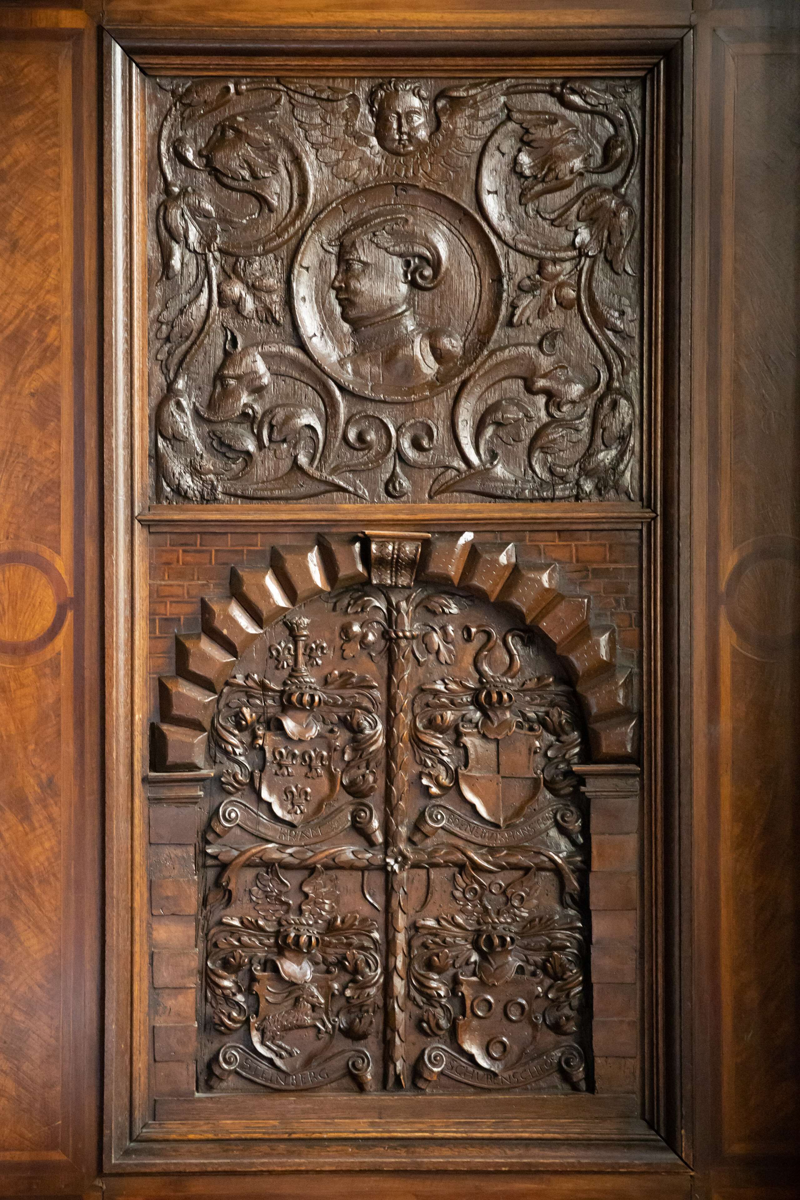 A detailed wooden door with intricate carvings, including a central profile portrait and heraldic designs.