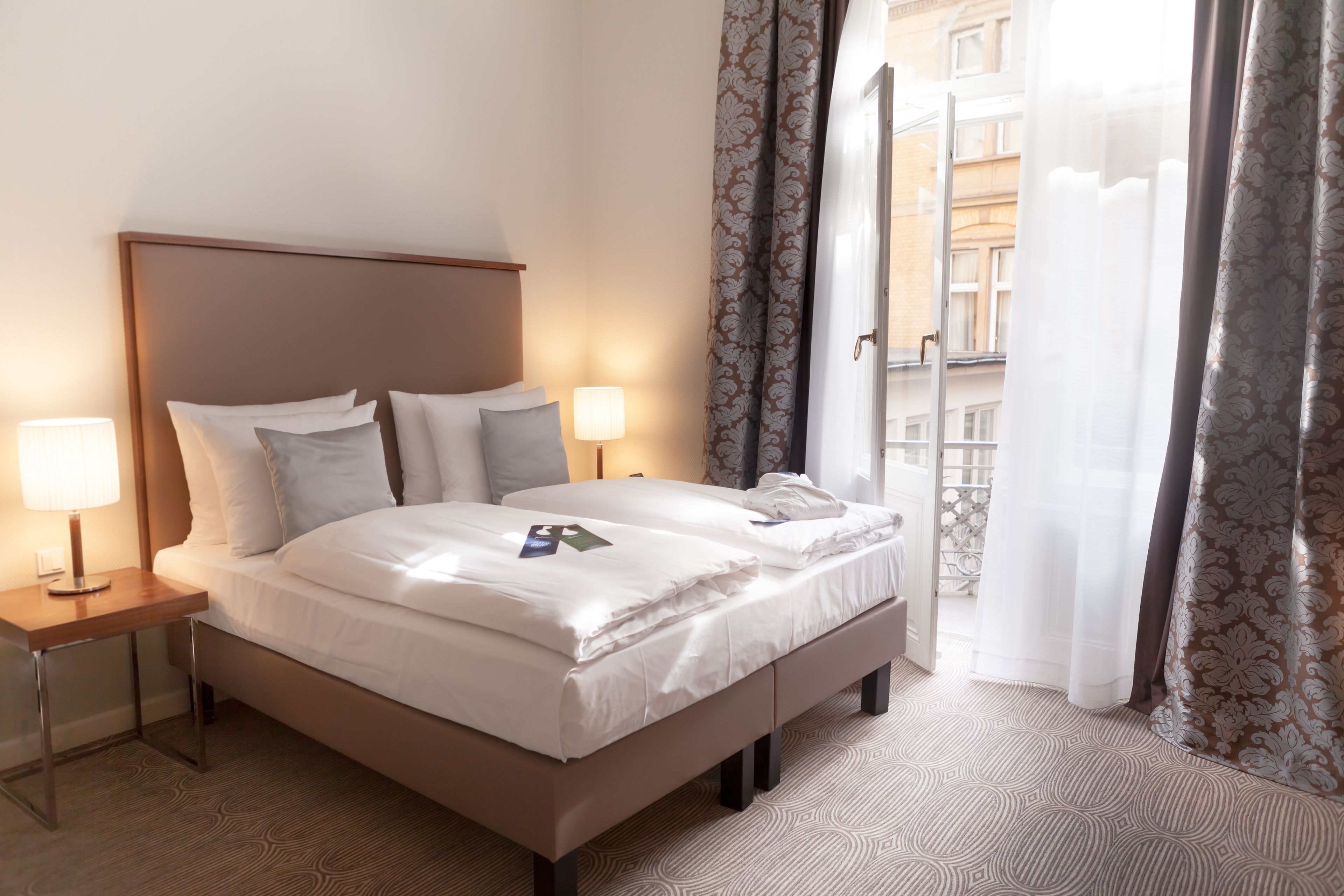 A neatly made bed with white linens, two bedside lamps, and a view of a building through sheer curtains.