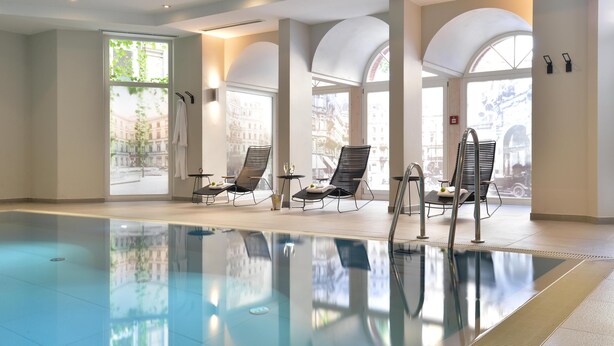 Una piscina con sillas y mesas de descanso, amplias ventanas en arco y vistas de edificios al exterior.