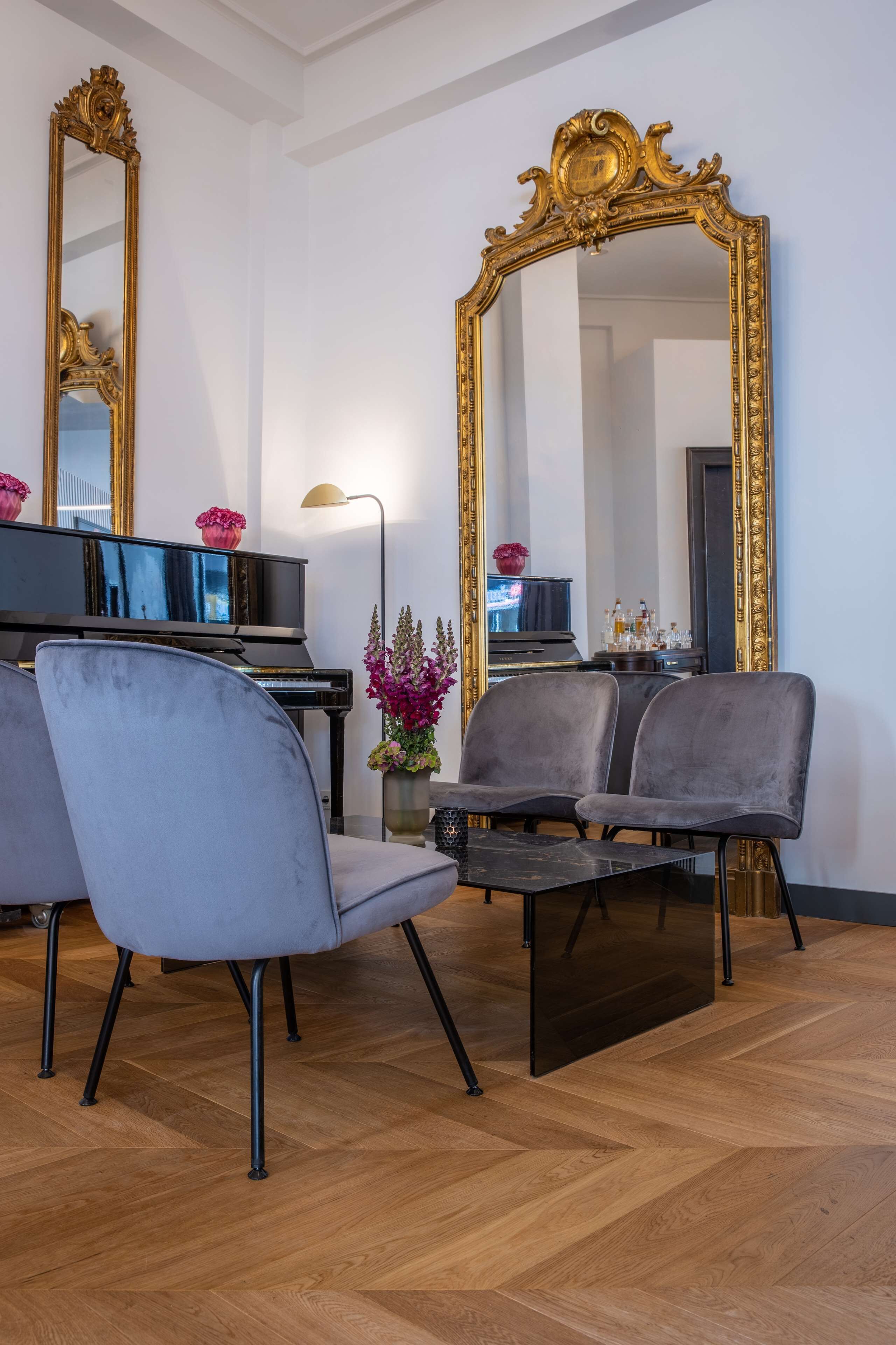 A living room with a grand piano, two grey upholstered chairs, a black coffee table, and a large ornate mirror.