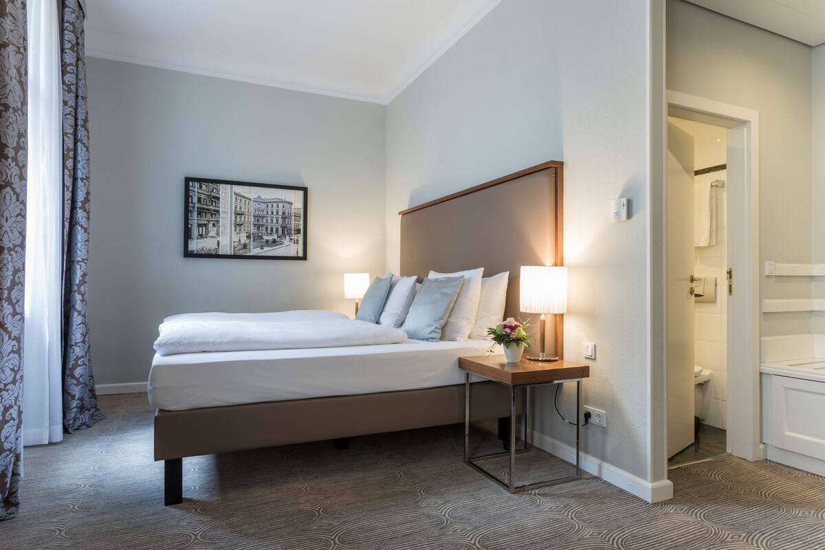 Une chambre d’hôtel bien rangée, avec un lit, une table de chevet, une lampe et un tableau encadré au mur.