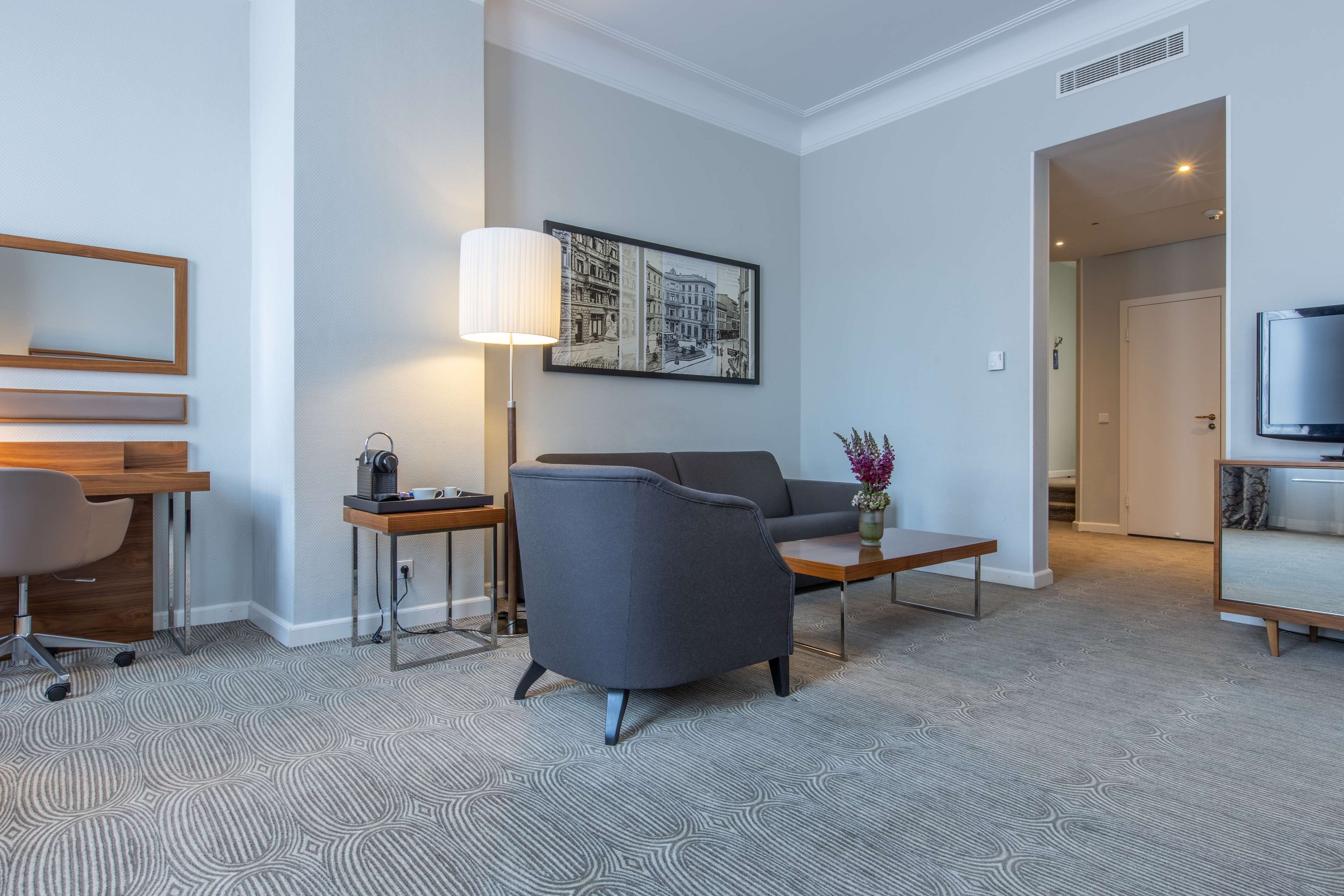Une chambre d’hôtel moderne avec un fauteuil gris, une petite table avec une lampe, un bureau et un téléviseur.