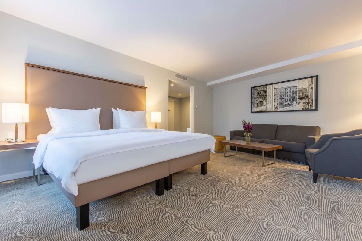 Une chambre d’hôtel avec un grand lit, un canapé, une table basse et un tableau représentant un paysage urbain accroché au mur.