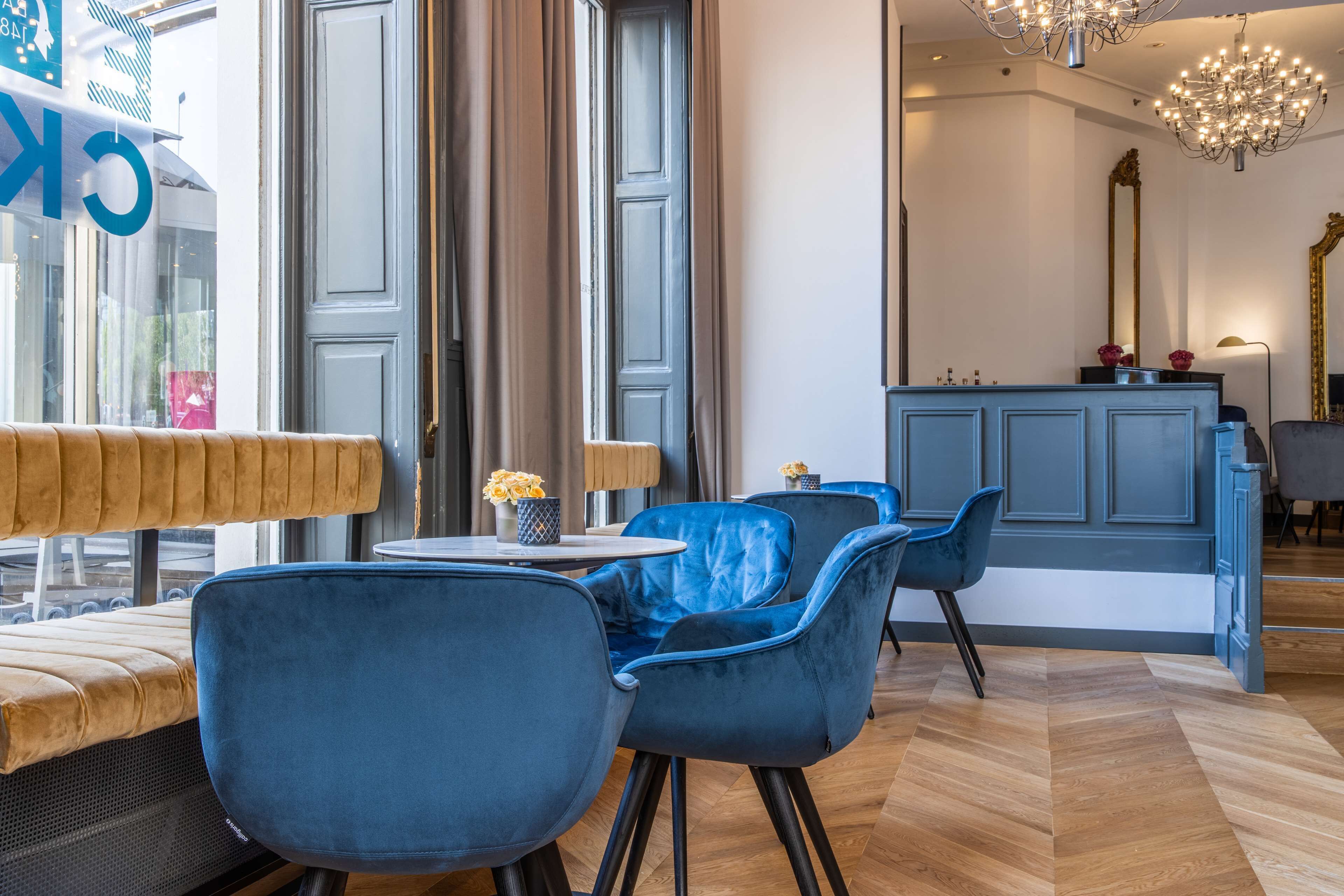 Un hall d’hôtel moderne avec des fauteuils en velours bleu, un banc en bois, une table ronde ornée d’un vase de fleurs et un coin bar avec un comptoir bleu.