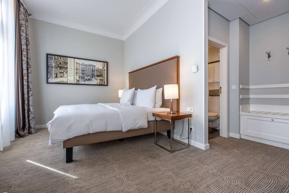 Une chambre d’hôtel avec un lit, une table de chevet, une lampe et un tableau représentant une vue de la ville, accroché au mur.