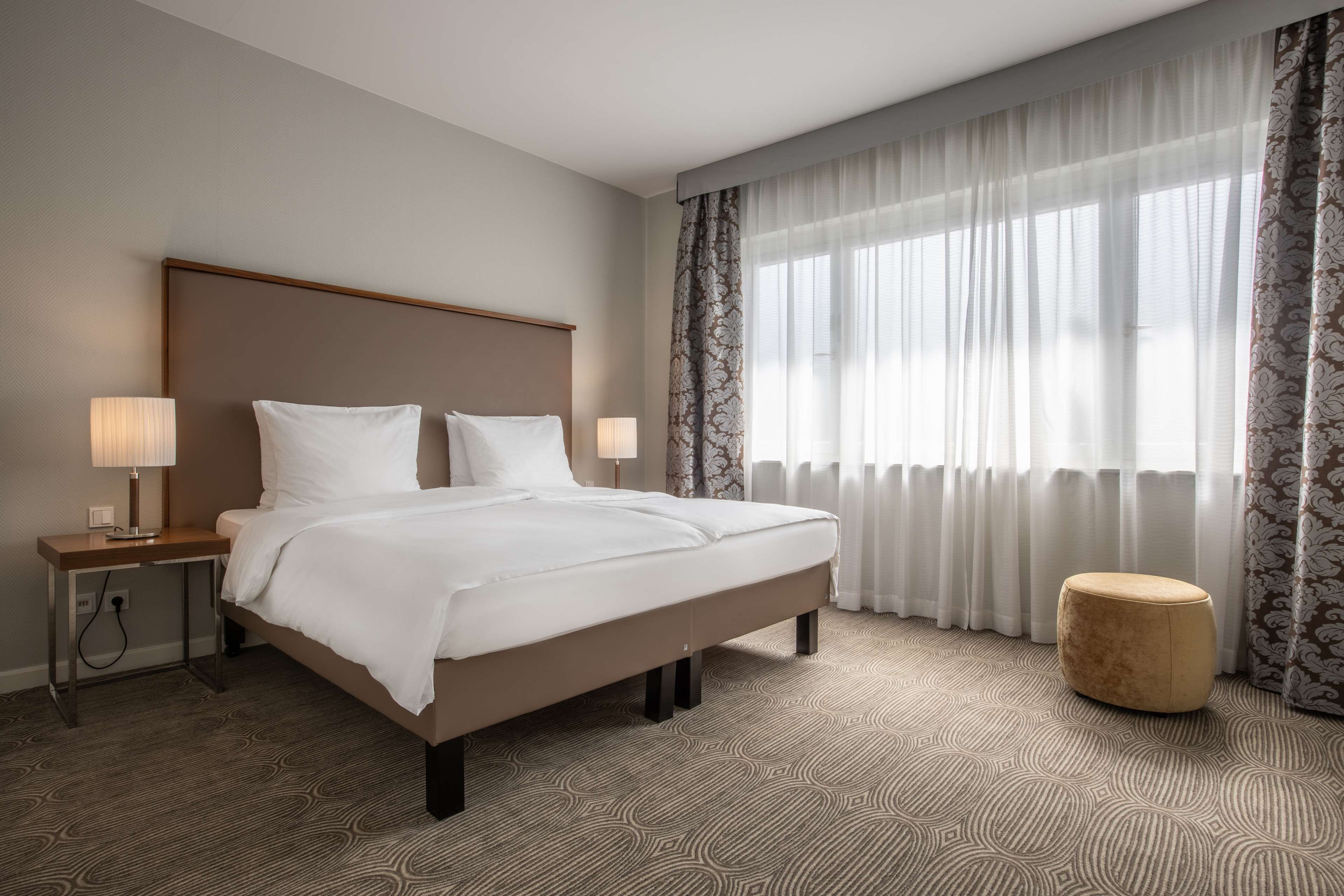 Una habitación de hotel con una cama grande, dos mesitas de noche con lámparas, un taburete redondo y un ventanal con cortinas translúcidas.