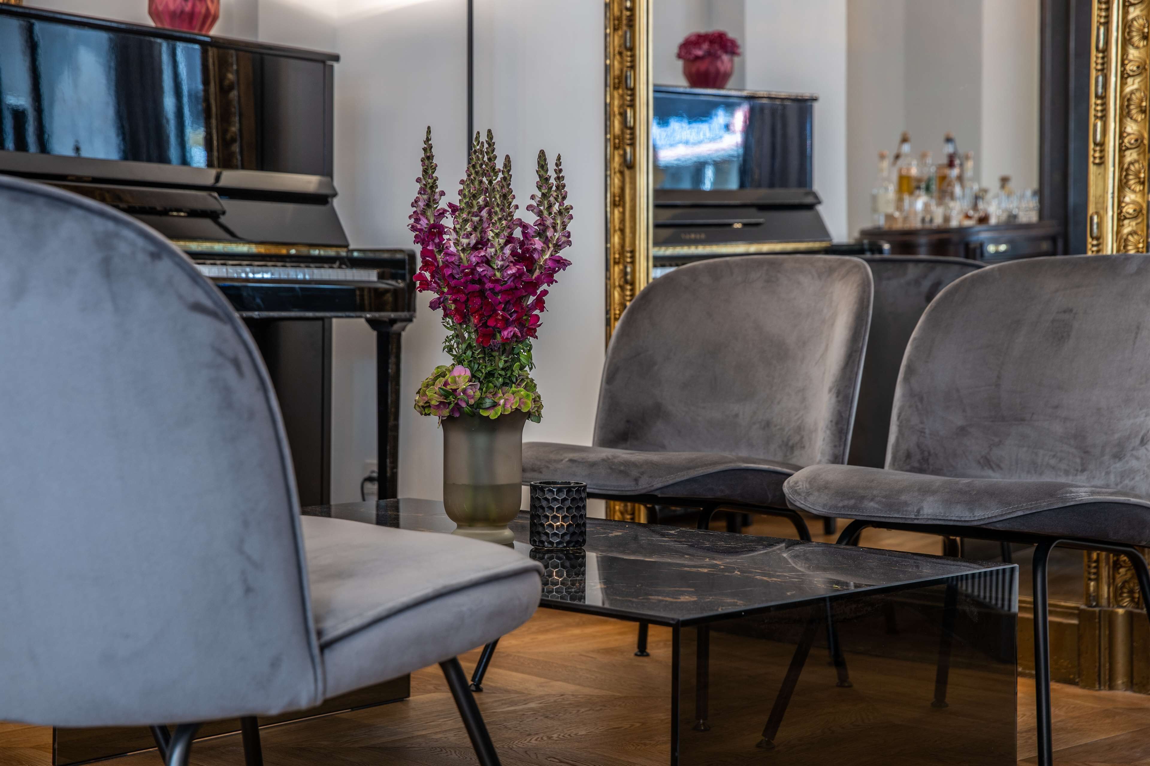 A modern living room with a piano, two grey upholstered chairs, a black coffee table, and a vase with purple flowers.