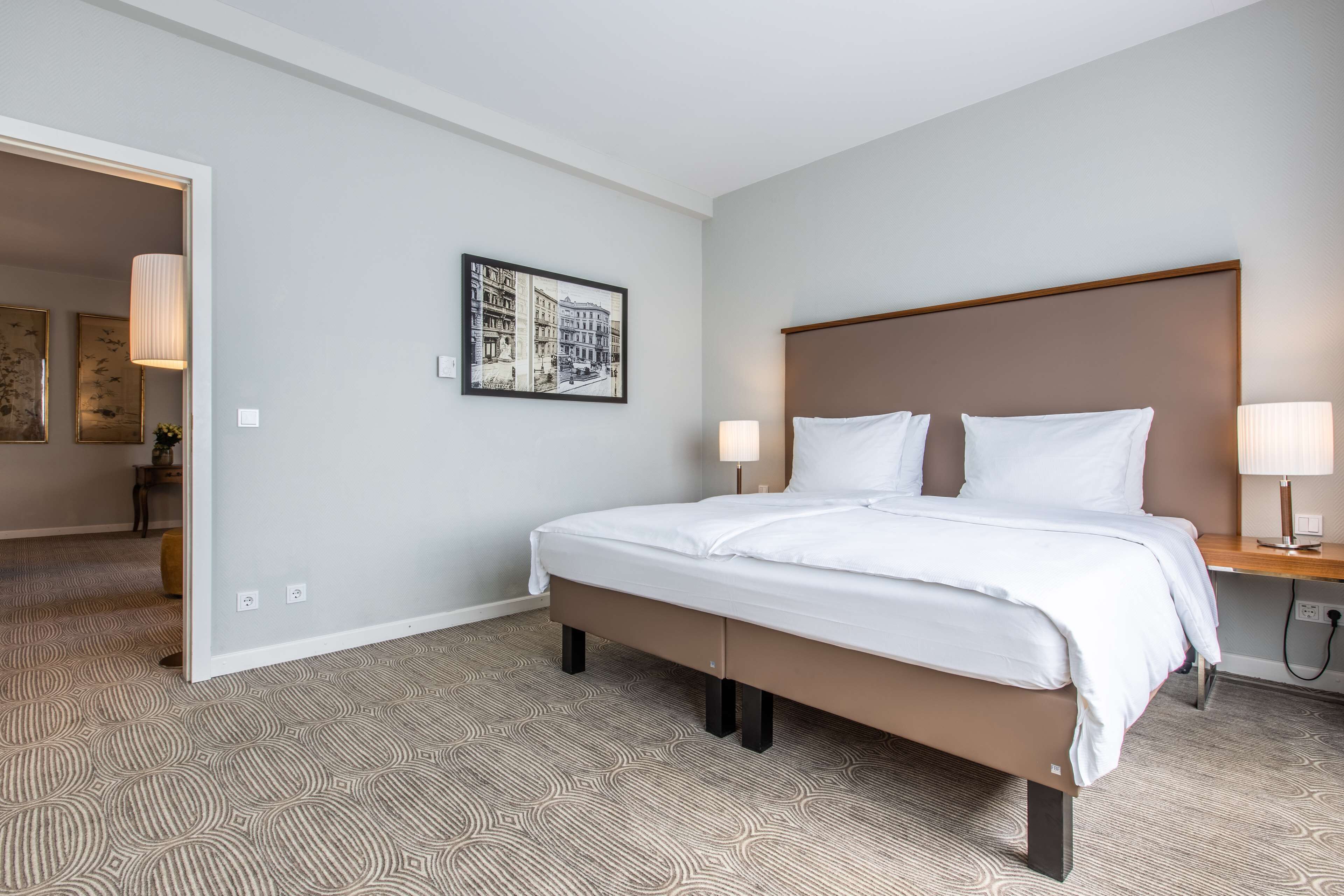 A hotel room with a large bed, a headboard, two bedside lamps, a wall-mounted picture, and a patterned carpet.