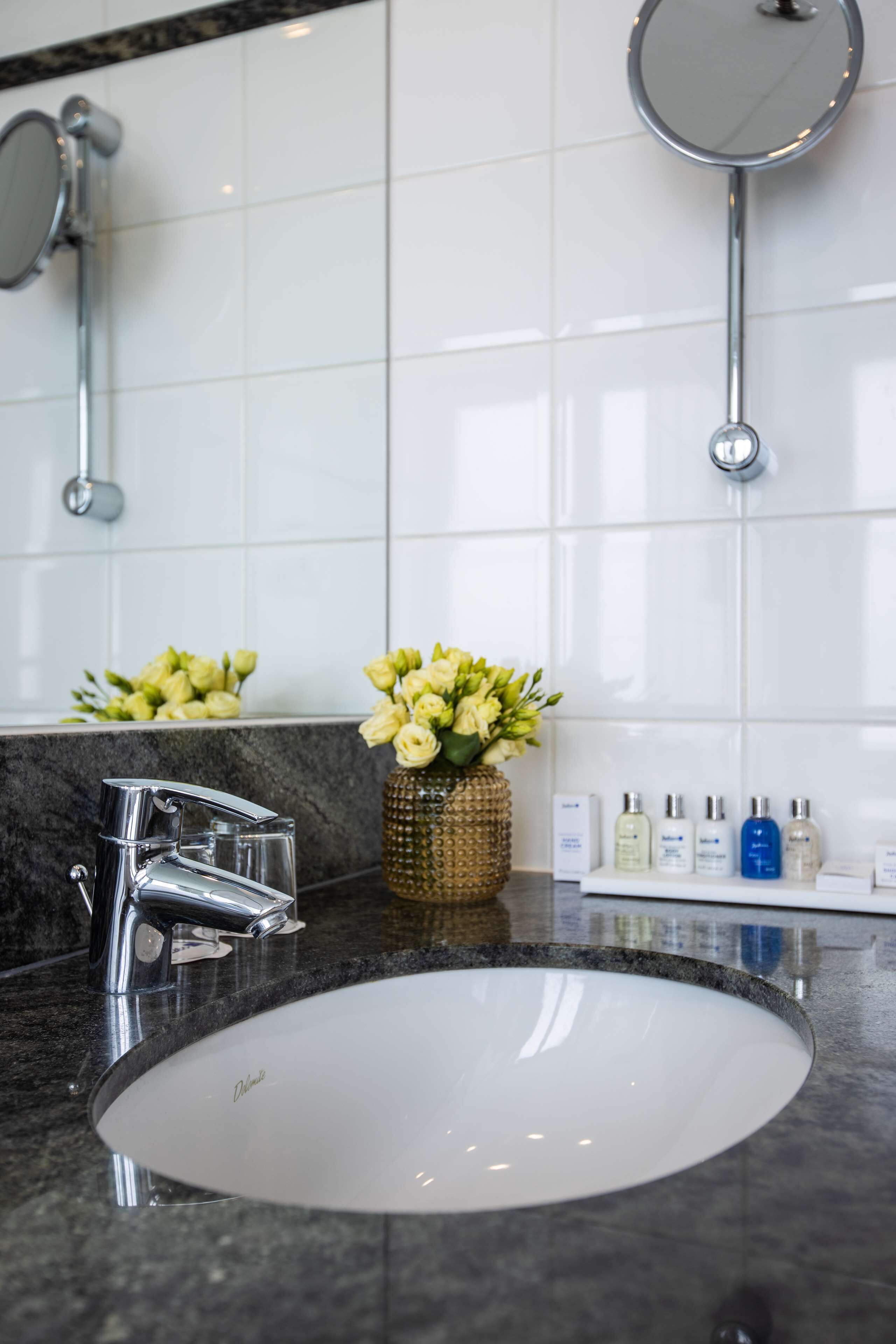 Un baño con un lavabo blanco, grifo cromado y un espejo encima. Hay flores en un jarrón sobre la encimera.