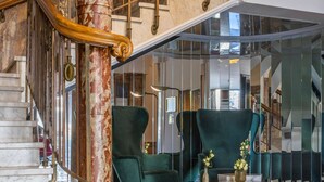 Una imponente escalera con una columna de mármol y barandilla de madera, que conduce a un interior moderno con sillones tapizados en verde y una mesa de centro de mármol.