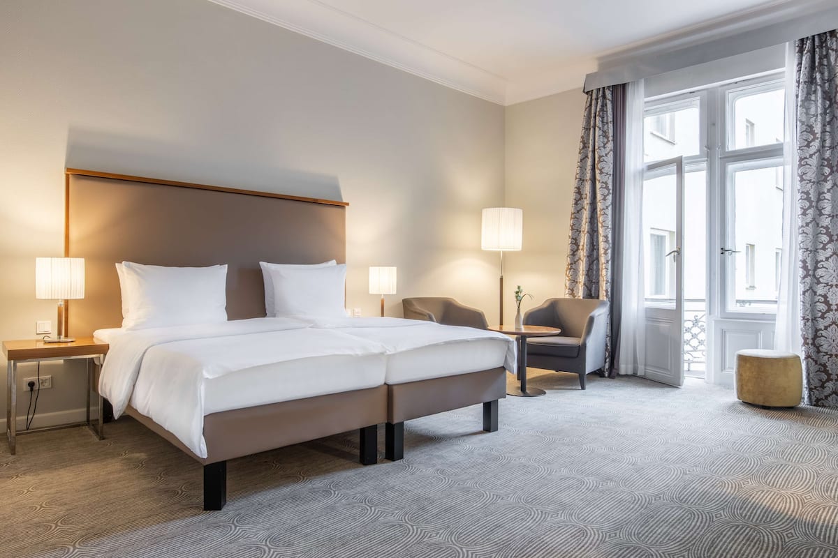 Une chambre d’hôtel avec un grand lit, deux tables de chevet avec des lampes, une chaise, un petit pouf et une fenêtre avec des rideaux.