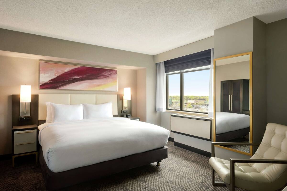 A hotel room with a large bed, two bedside tables with lamps, a chair, a large window with a view, and a painting on the wall.