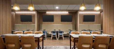 Un salón de conferencias con mesas, sillas, dos pantallas grandes e iluminación colgante.