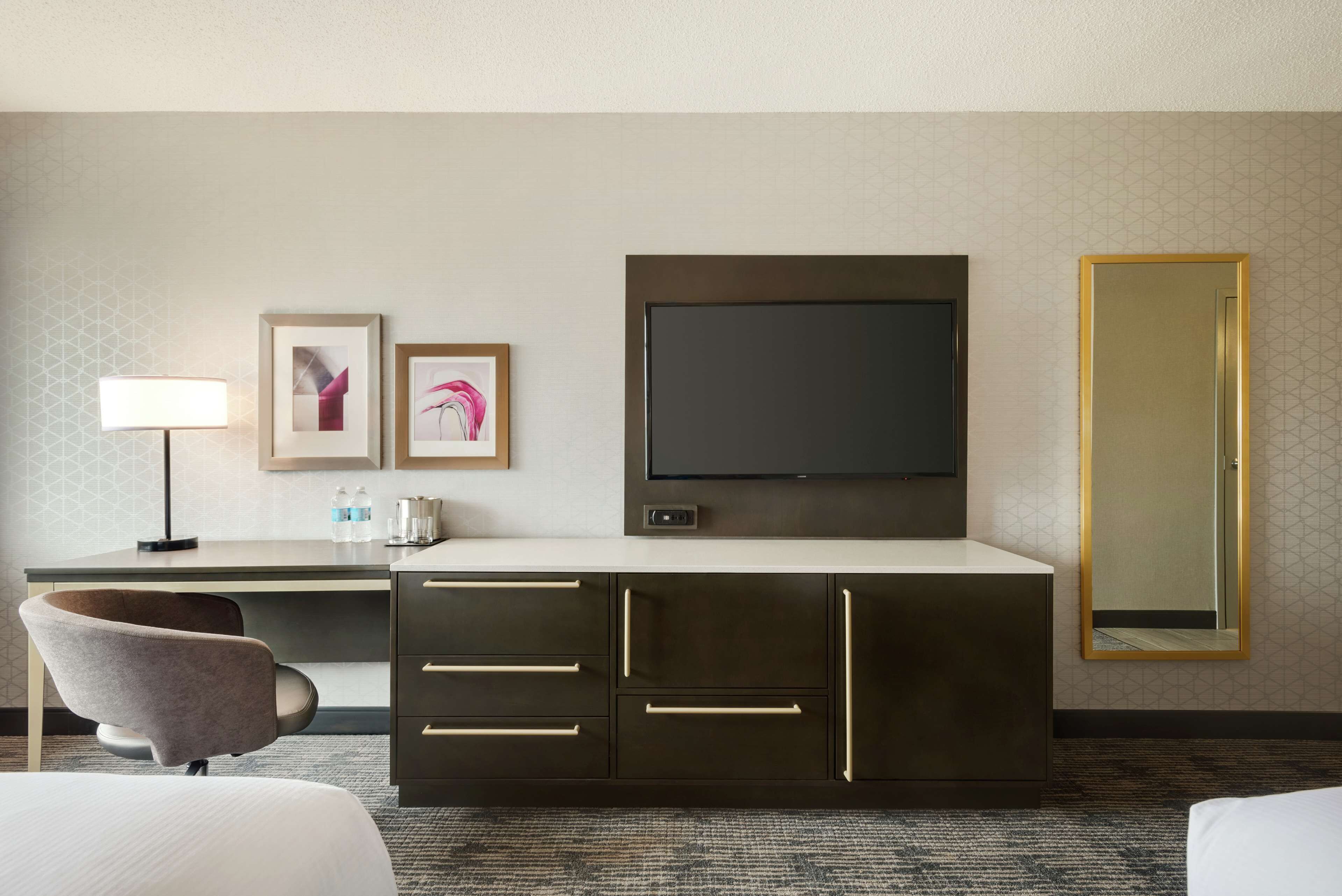 Una habitación de hotel con cama, escritorio con lámpara, televisor y un espejo grande.