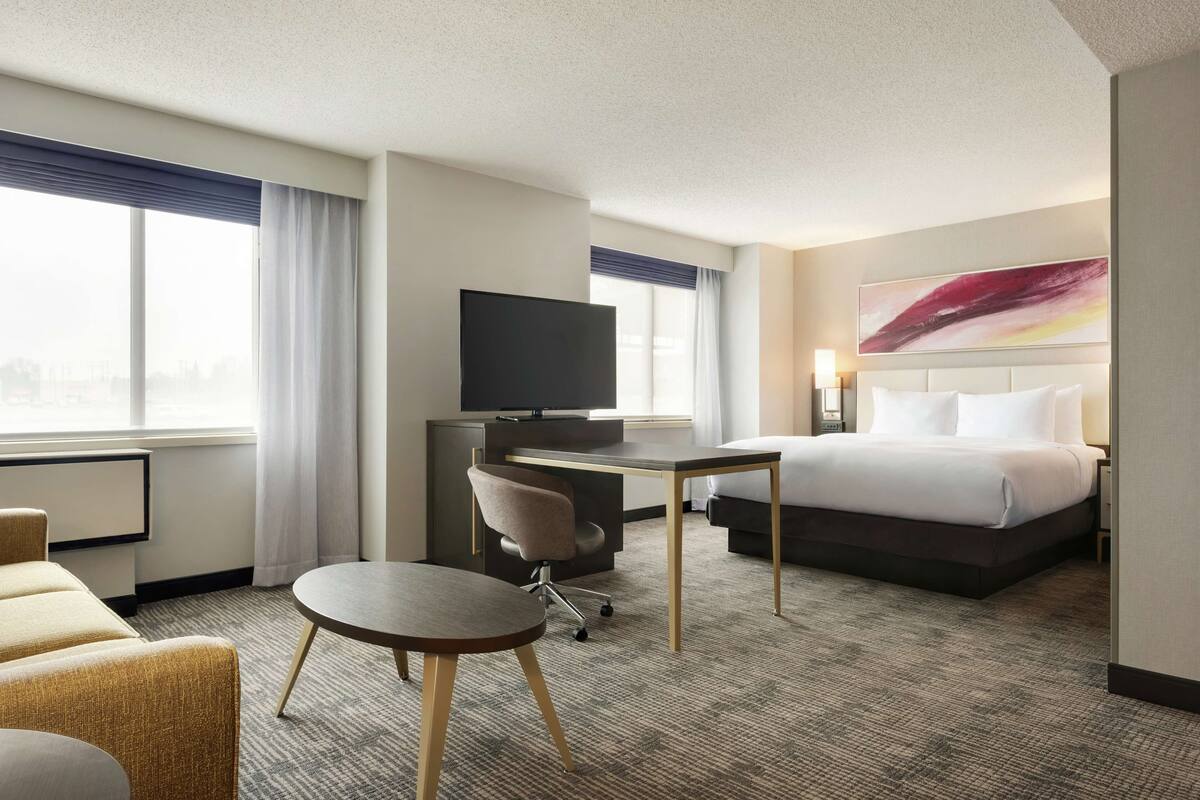 A hotel room with a bed, a desk with a chair, a television, and a large window with curtains.