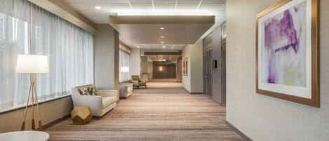 A hotel corridor with a carpeted floor, a sofa, a lamp, and a painting on the wall.