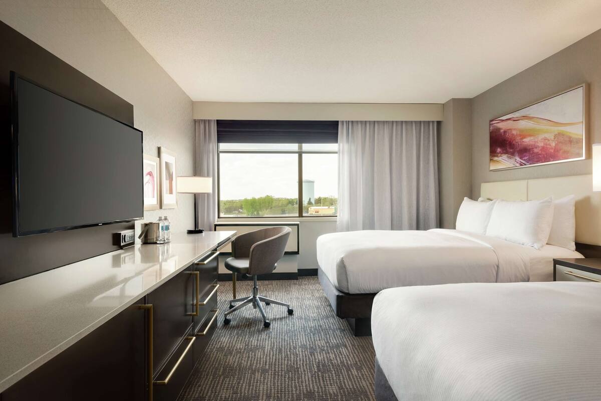 A hotel room with a flat-screen TV, a desk with a chair, two beds, a large window with curtains, and a painting on the wall.