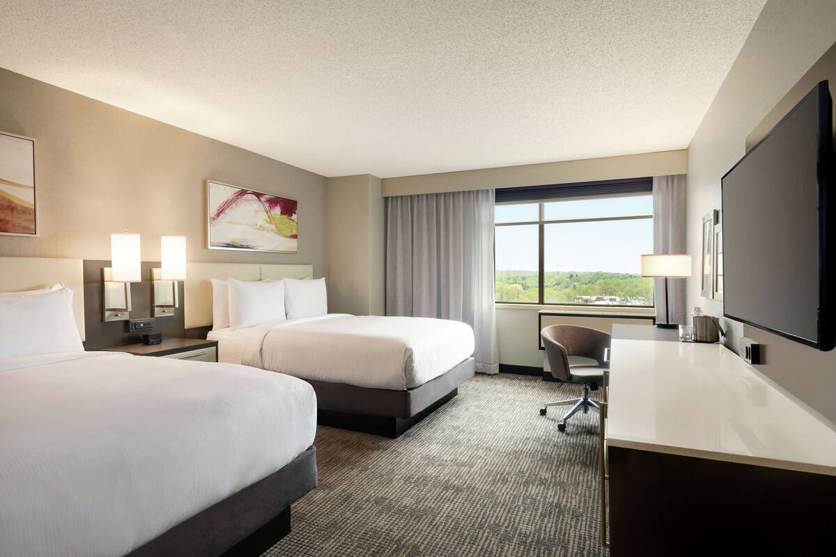 A hotel room with two beds, a desk, a chair, a TV, and a large window with a view.