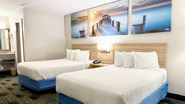 A hotel room with two beds, a wooden headboard, a nightstand, a lamp, and a painting of a pier on the wall.