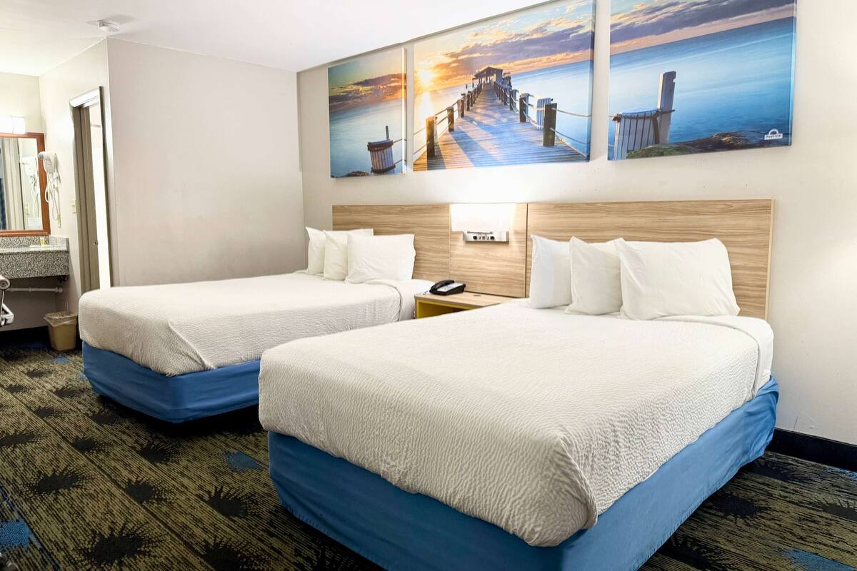 A hotel room with two beds, a wooden headboard, a nightstand, a lamp, and a painting of a pier on the wall.