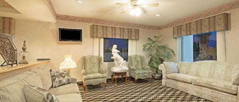 A living room with patterned carpet, two armchairs, a sofa, a ceiling fan, and a television on the wall.